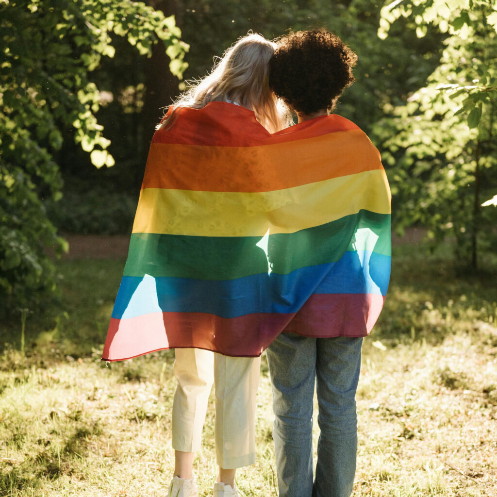 Dos personas de aspaldas que se abrazan y tienen una bandera arcoiris sobre sus espaldas